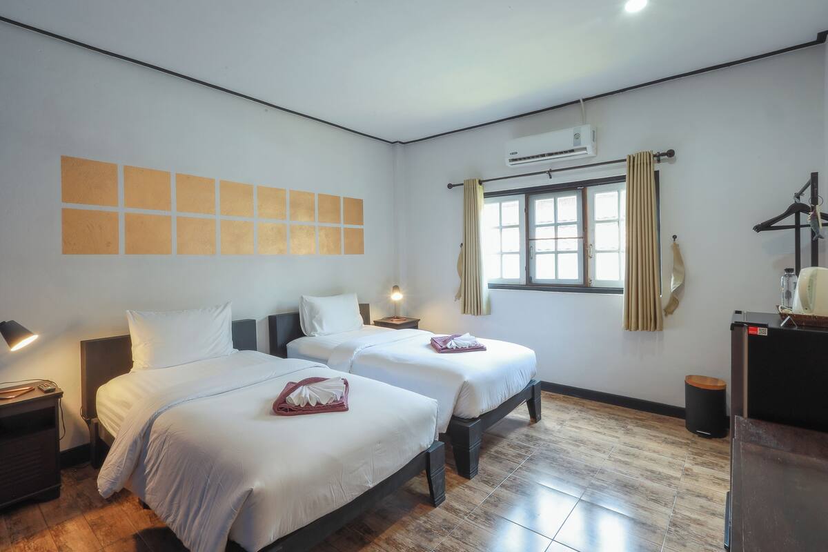 Habitación de hotel con dos camas, una ventana con cortinas, un equipo de aire acondicionado y una pequeña mesa con lámpara.