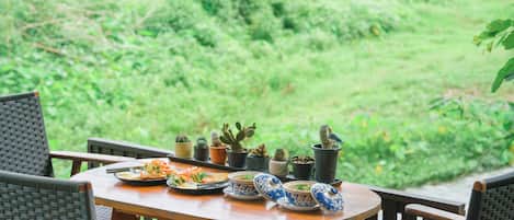 Une table en bois, sur laquelle sont disposés des plats de nourriture et des bols aux motifs bleus et blancs, entourée de chaises en osier, avec des cactus en pot en arrière-plan.