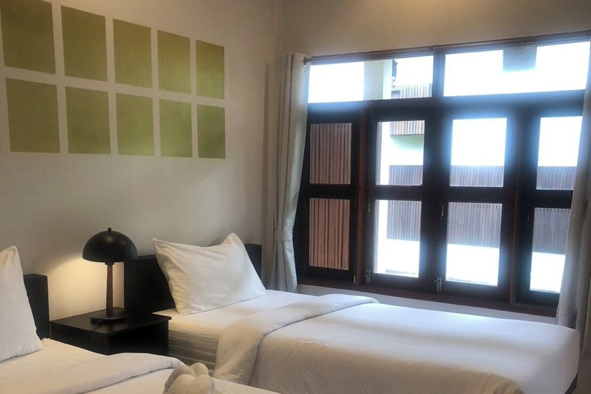 Habitación de hotel con dos camas, una mesita de noche con lámpara, ventana con cortinas y pared con un diseño geométrico.
