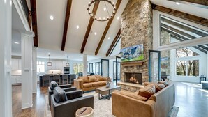 A spacious living area with a stone fireplace, leather sofas, and a large window overlooking a forest. - Comfy Bear Mountain Lodge| Family-Friendly (Helen)