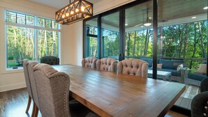 A dining area with a long wooden table, upholstered chairs, and large windows offering a view of trees. - Comfy Bear Mountain Lodge| Family-Friendly (Helen)