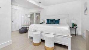 Un dormitorio moderno con una cama blanca, dos taburetes redondos, un sofá gris y una ventana con persianas.