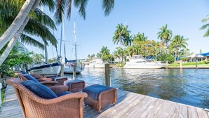 A wooden dock with wicker furniture and a view of boats on a calm water body, surrounded by palm trees. - The Riverside Retreat #2 (Fort Lauderdale)