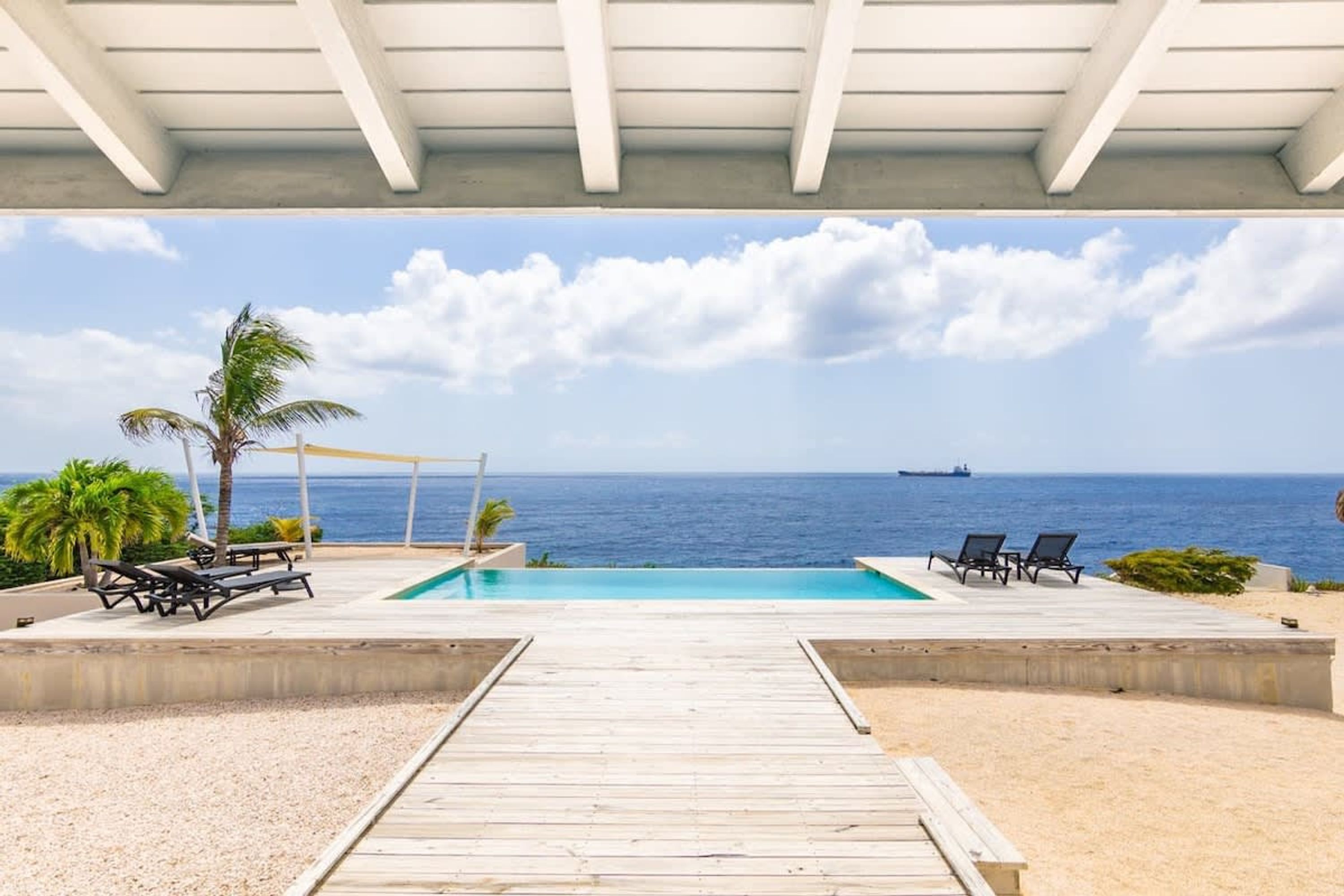 Um deck de madeira leva a uma piscina com espreguiçadeiras e palmeiras, com vista para o oceano.