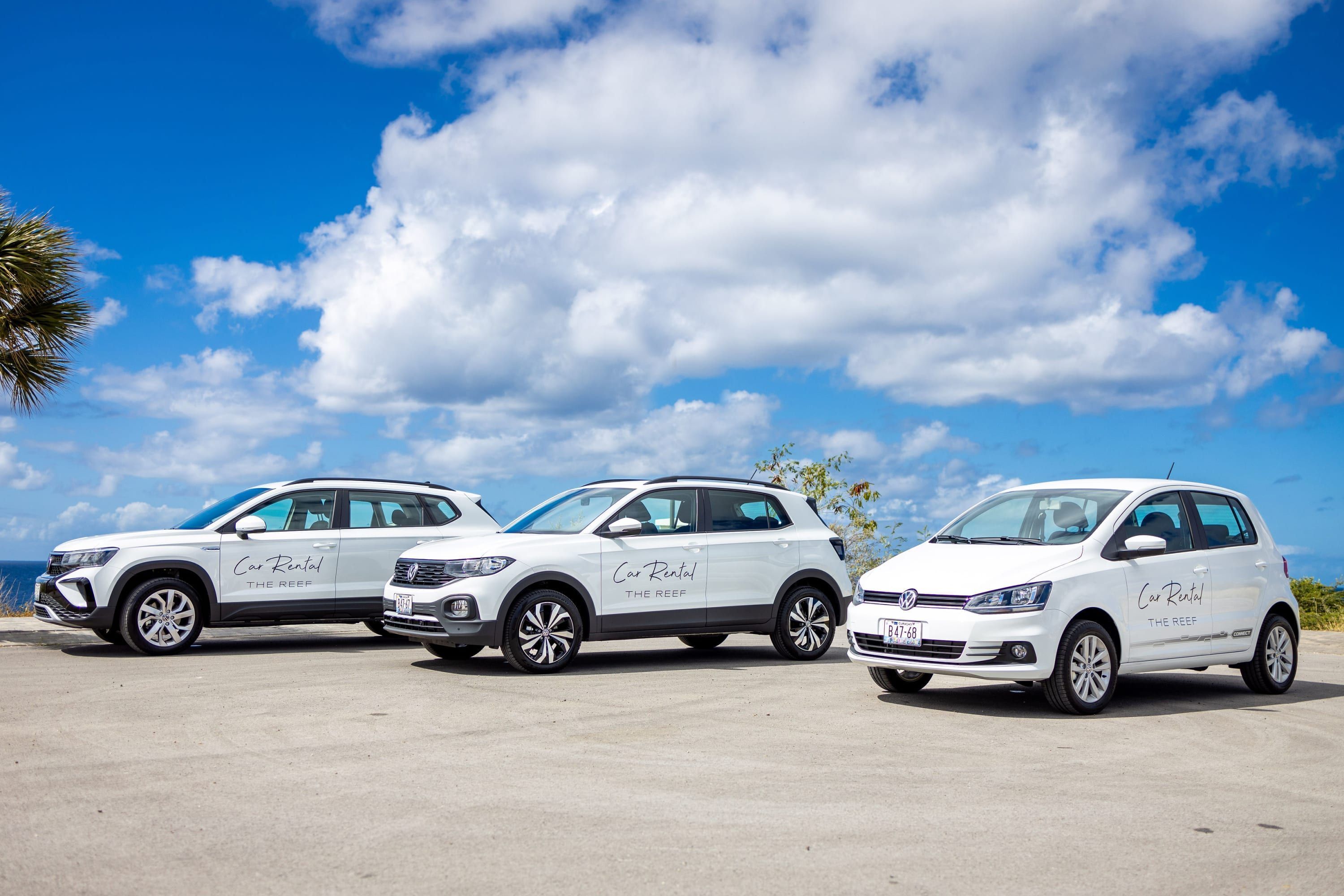 Three white cars parked on a concrete surface with a clear blue sky and clouds in the background.