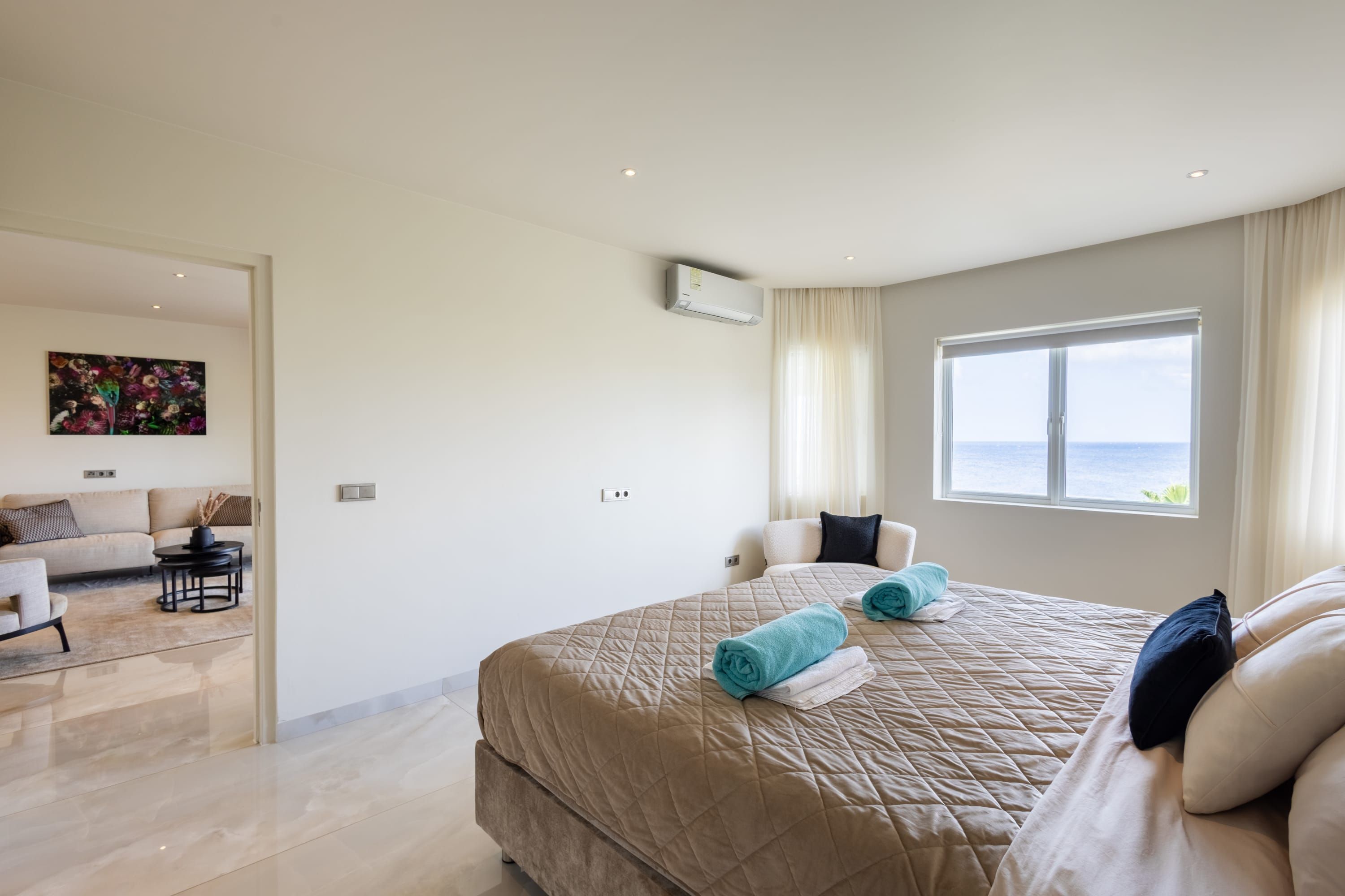 A modern hotel room with a large bed, a seating area with a sofa and armchairs, a painting on the wall, and a window with a view.