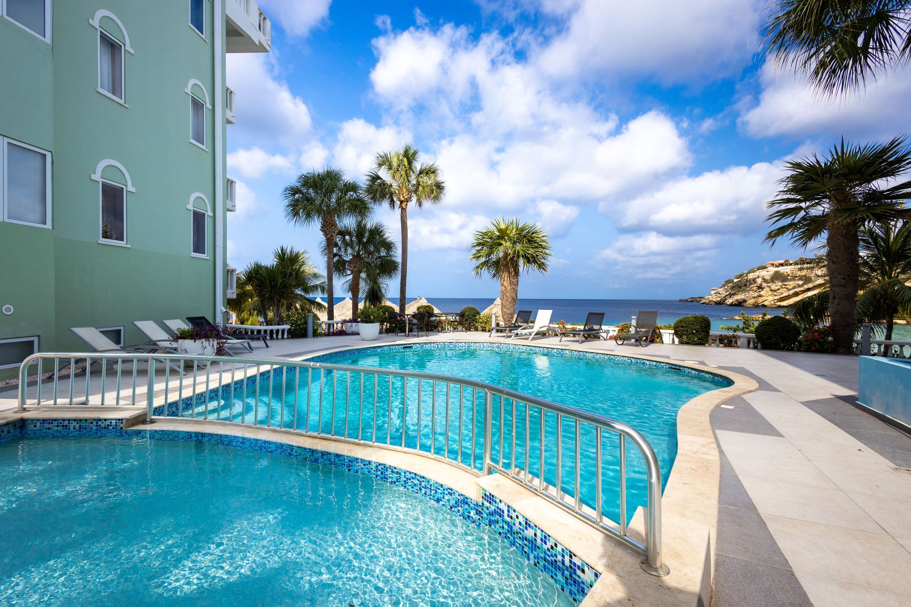 Ein Pool mit gewölbter Kante, umgeben von Liegestühlen und Palmen, mit einem Gebäude und Blick auf das Meer im Hintergrund.