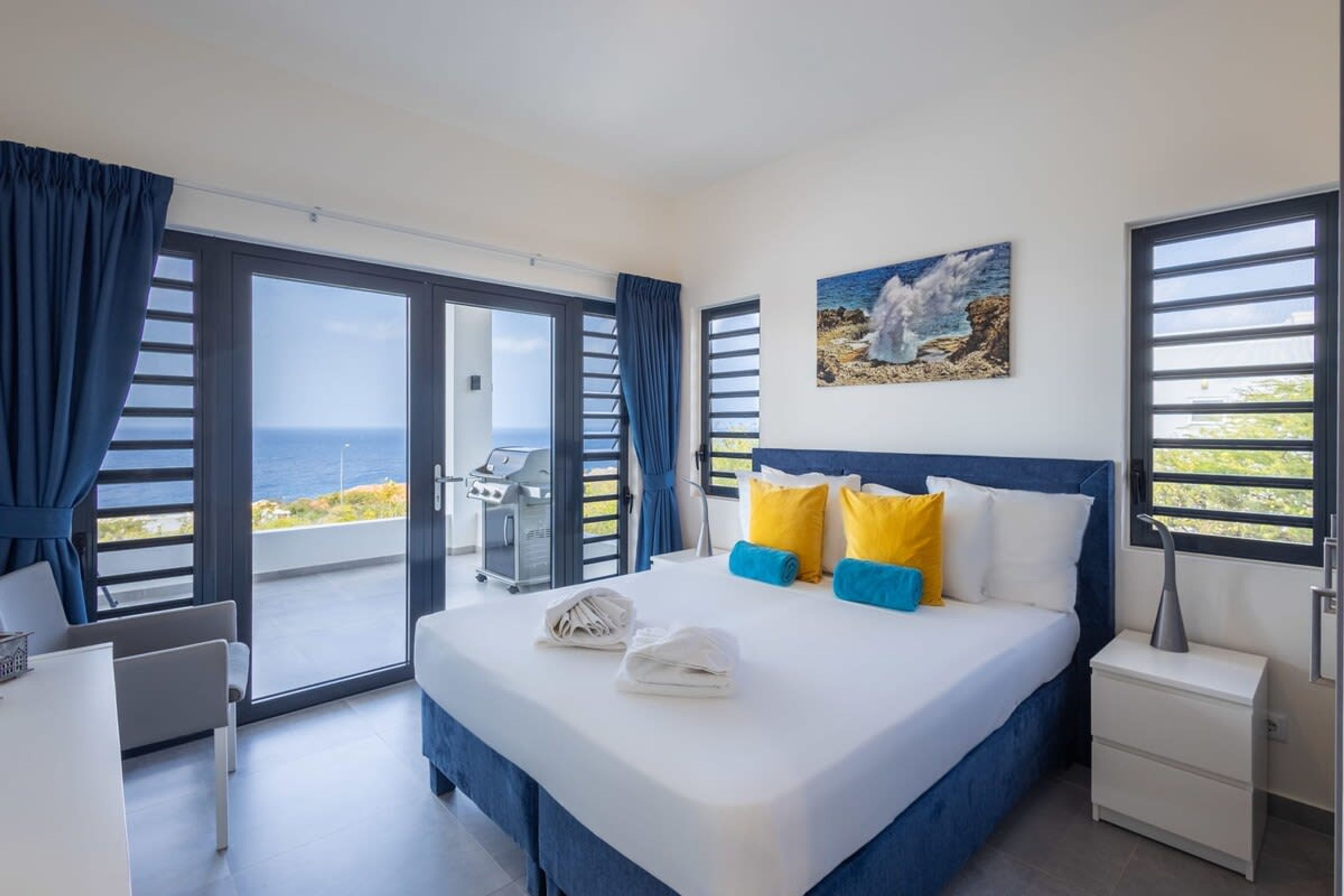 A bedroom with a bed, a desk, a chair, a nightstand, and a view of the sea.