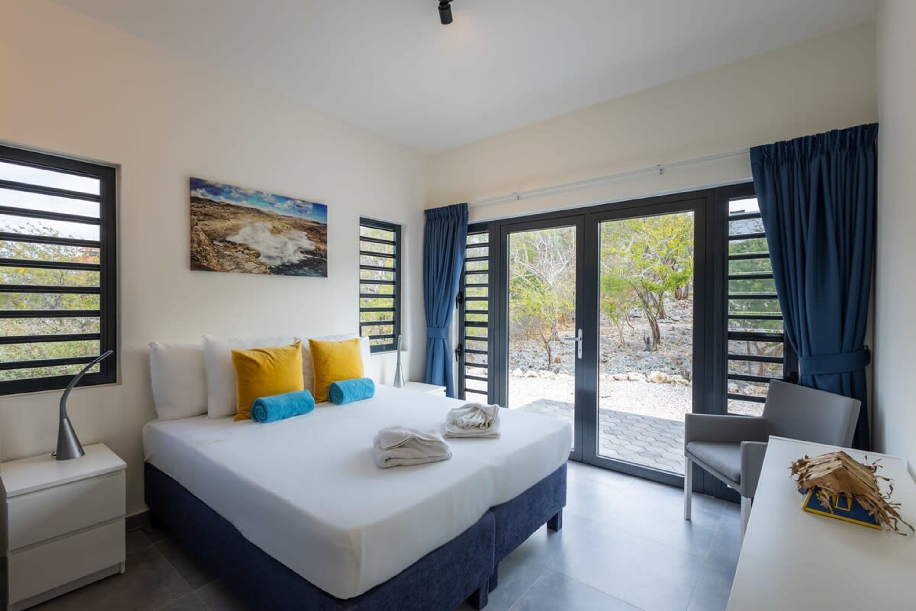 Quarto com uma cama, uma escrivaninha e vista pela porta de vidro.
