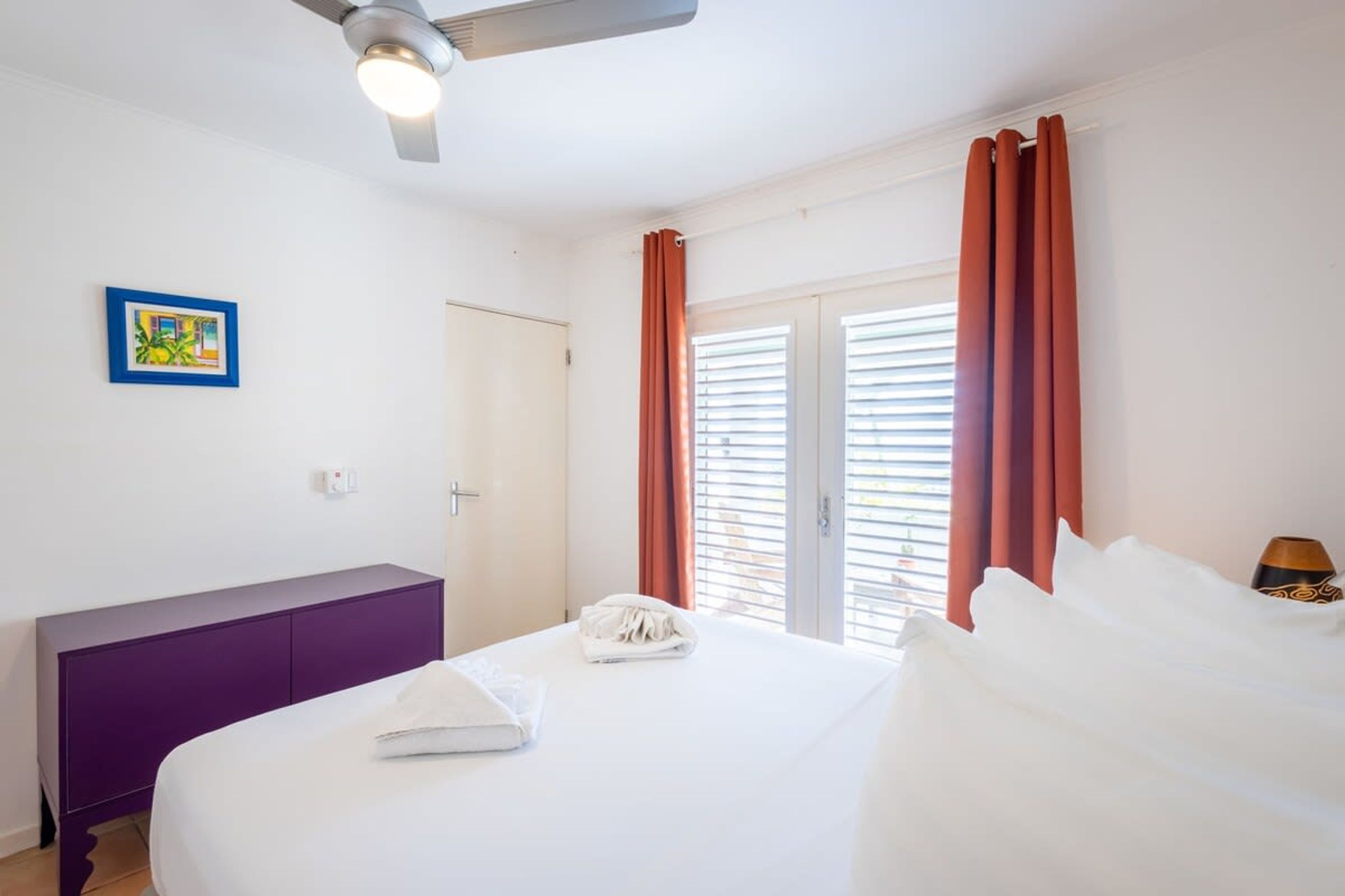 A hotel room with a bed, a purple dresser, a ceiling fan, and a window with orange curtains.