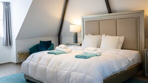 Un letto rifatto con lenzuola bianche e color petrolio, una lampada da comodino e una poltrona color petrolio in una stanza con soffitto spiovente e finestra con tende.