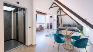 Un interno con un ascensore, zona pranzo con tavolo in vetro e sedie color acquamarina, e una cucina sullo sfondo.