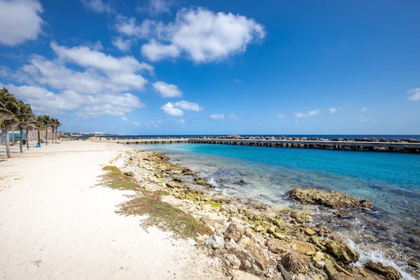 Uma praia com águas cristalinas, costa rochosa e um cais ao longe.