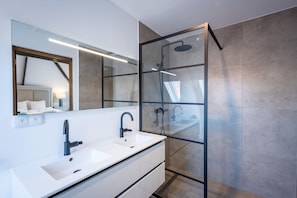 Uma casa de banho moderna com chuveiro em cabine de vidro, lavatório branco com torneira preta e um espelho grande.