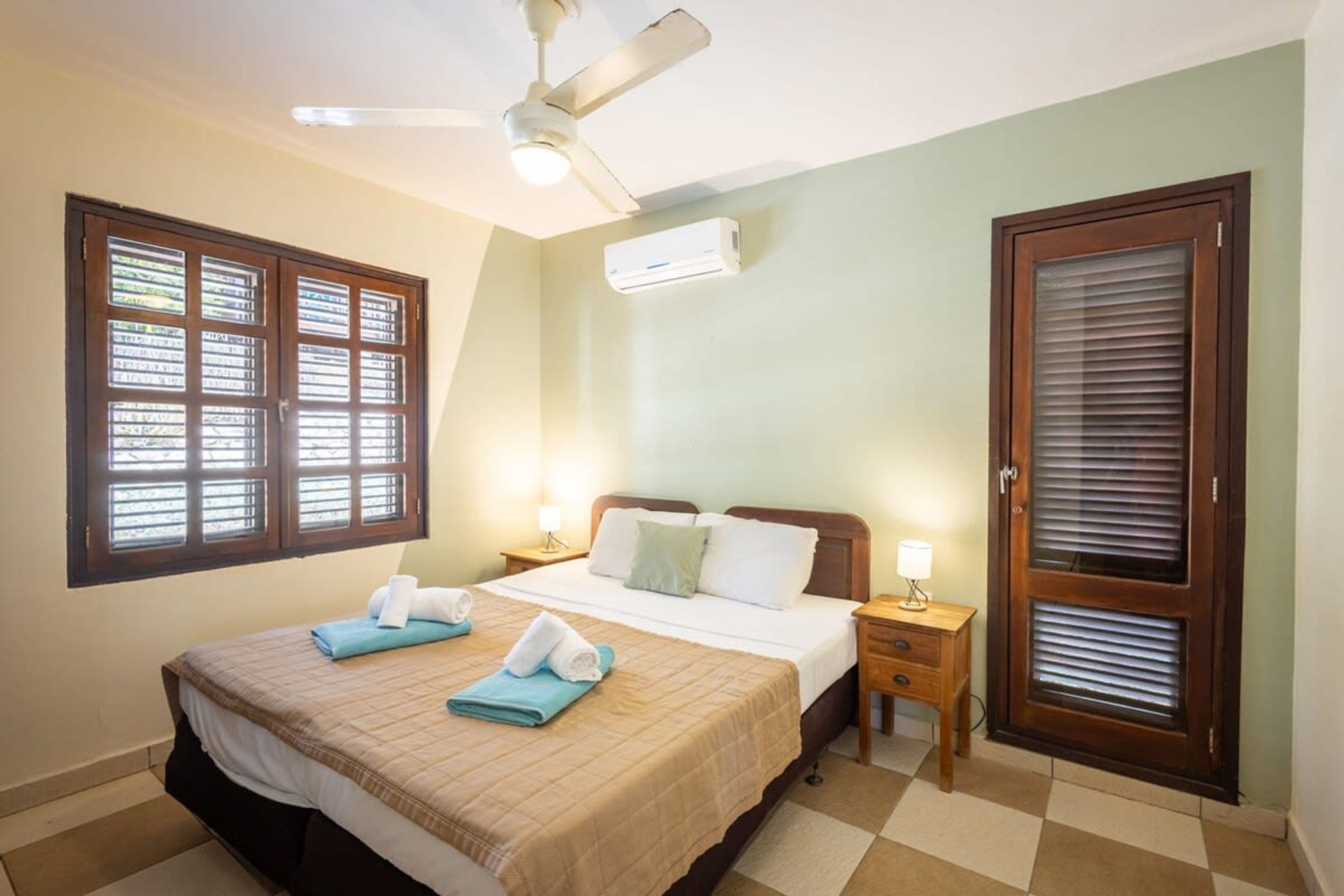 Habitación de hotel con cama, ventilador de techo, equipo de aire acondicionado, ventana con persianas, puerta con ventanal y mesita de noche con lámpara.