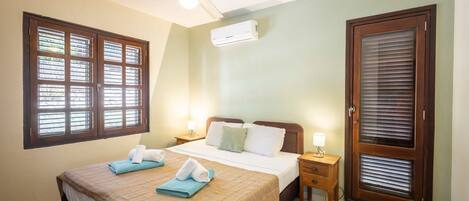 A hotel room with a bed, a ceiling fan, an air conditioning unit, a window with blinds, a door with a window, and a nightstand with a lamp.