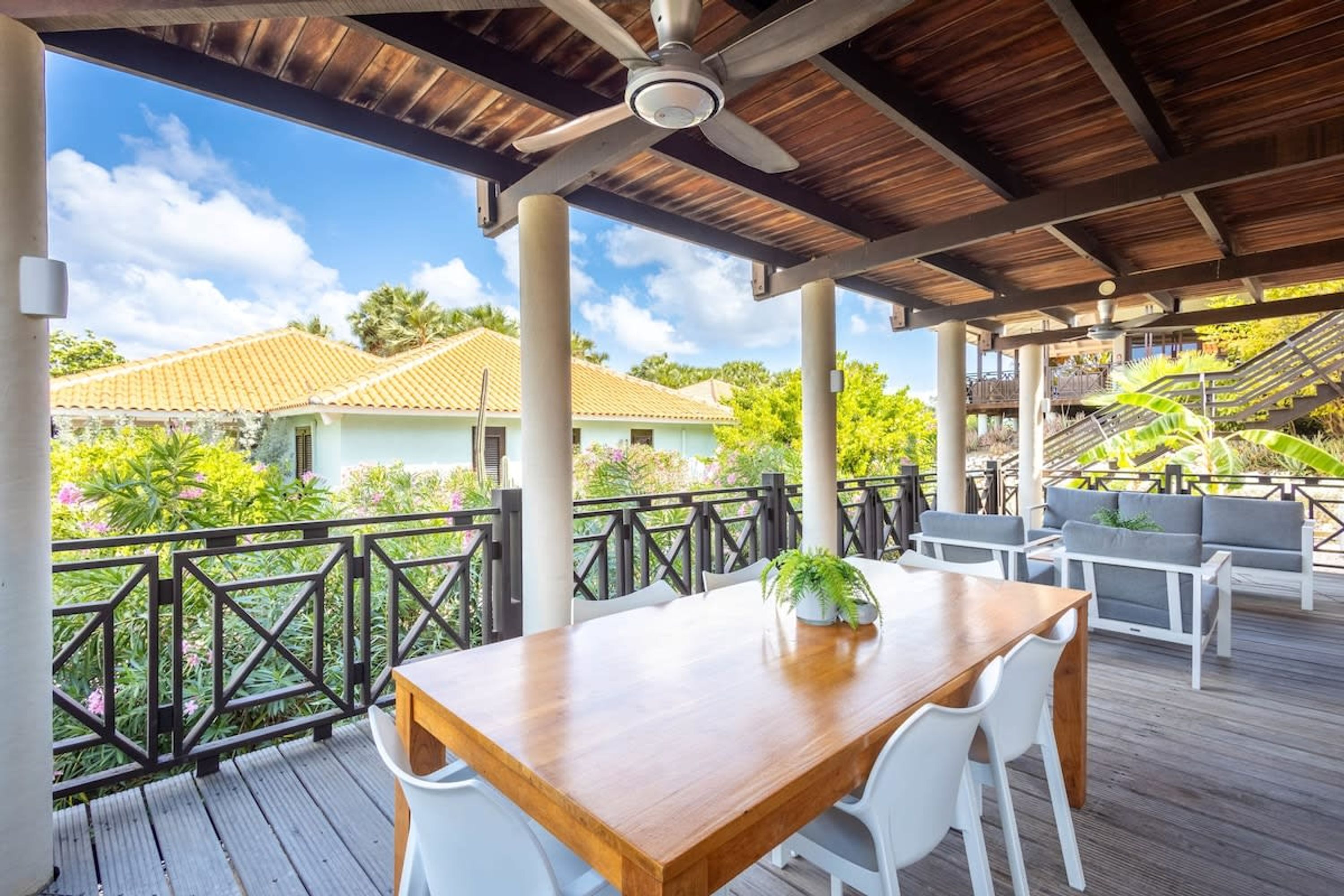 Una terraza de madera con una mesa y sillas, un ventilador de techo y una barandilla con vistas a un jardín y edificios.