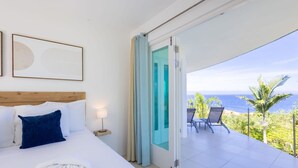 Habitaci ón de hotel con cama, mesita de noche, ventana con cortinas y balcón con vista al océano.