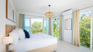 Un dormitorio amplio con una cama grande, una mesita de noche, una lámpara colgante y vistas a la vegetación a través de amplias ventanas.