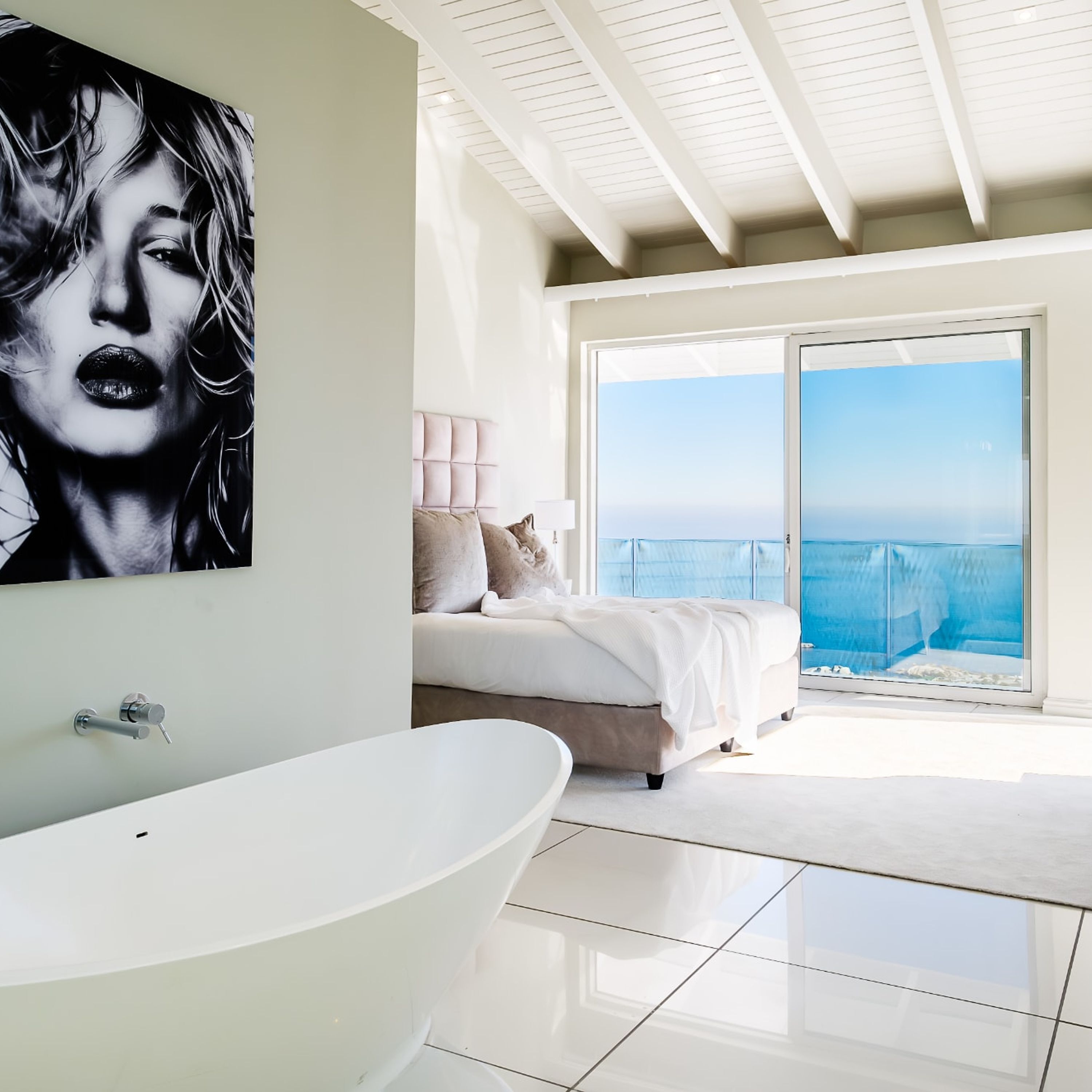 A modern bedroom with a large bed, a white bathtub, and a view of the ocean through a sliding glass door.