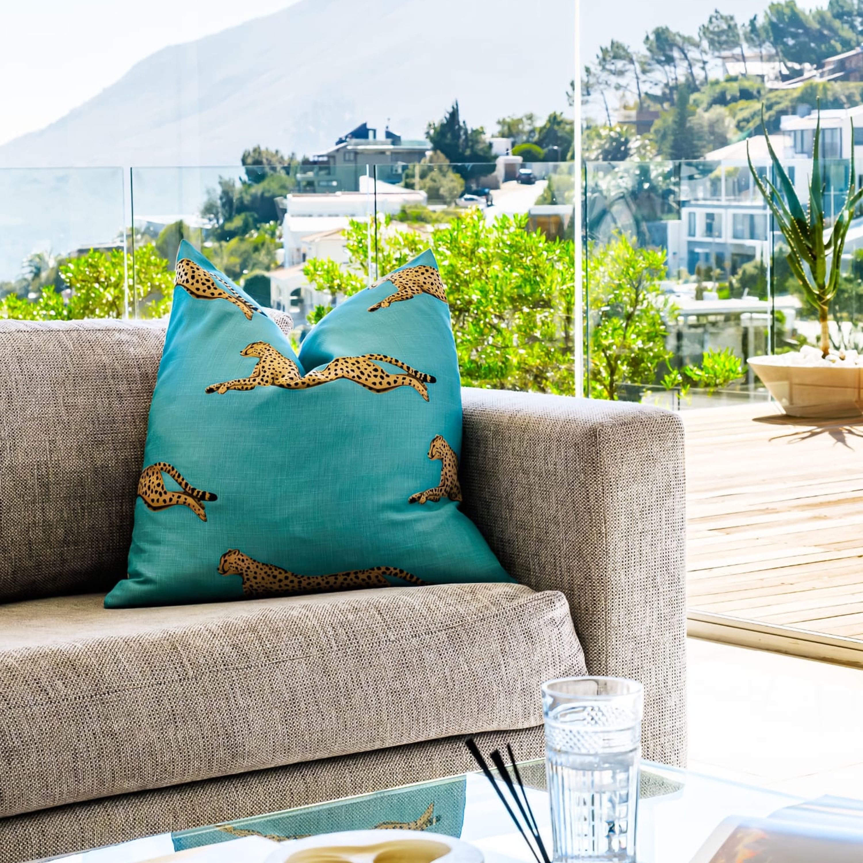 A sofa with teal cushions featuring cheetah prints, a glass table with a clear glass and a book, and a view of buildings and greenery in the background.