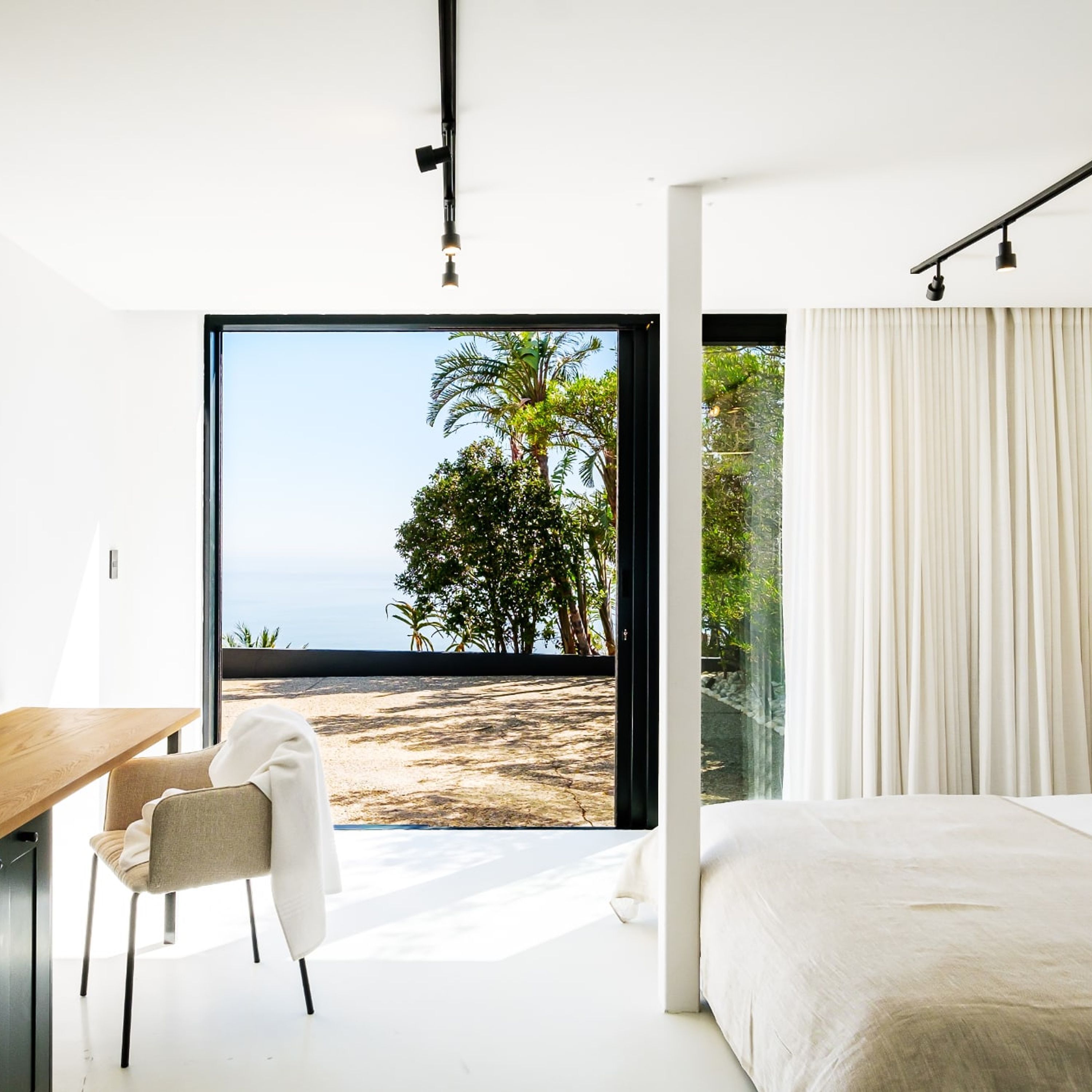 Un dormitorio moderno con una cama grande, un escritorio y una silla. Cuenta con un amplio ventanal que da a vista de playa.