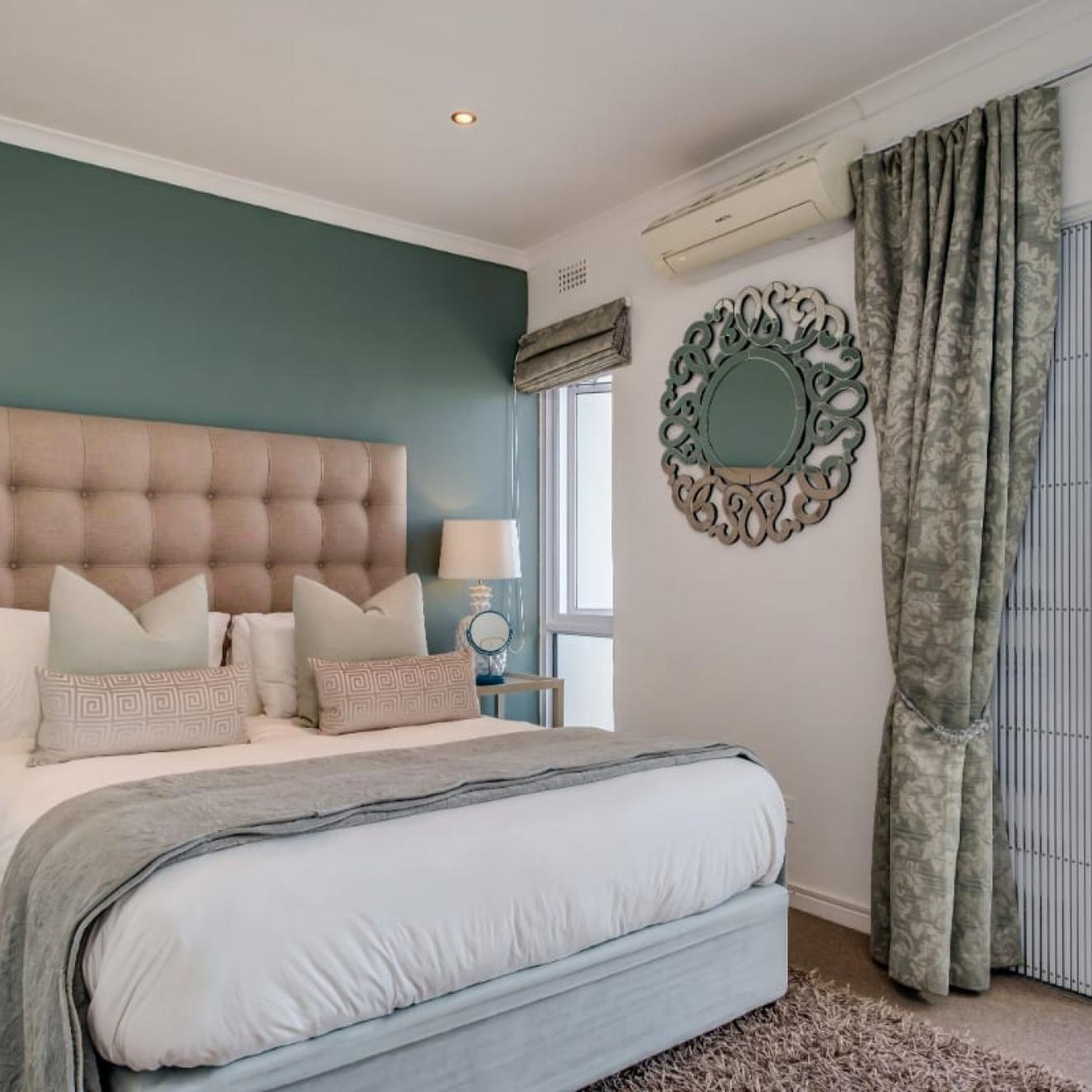 A bedroom with a tufted headboard, a bed with multiple pillows, a bedside table with a lamp, a decorative mirror, and a window with curtains.