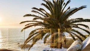 Een balkon met een palmboom, een lounge stoel en uitzicht op de oceaan bij zonsondergang.