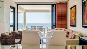 Un salon moderne avec une table basse en verre, des meubles en osier, un canapé beige et une vue sur l’océan.