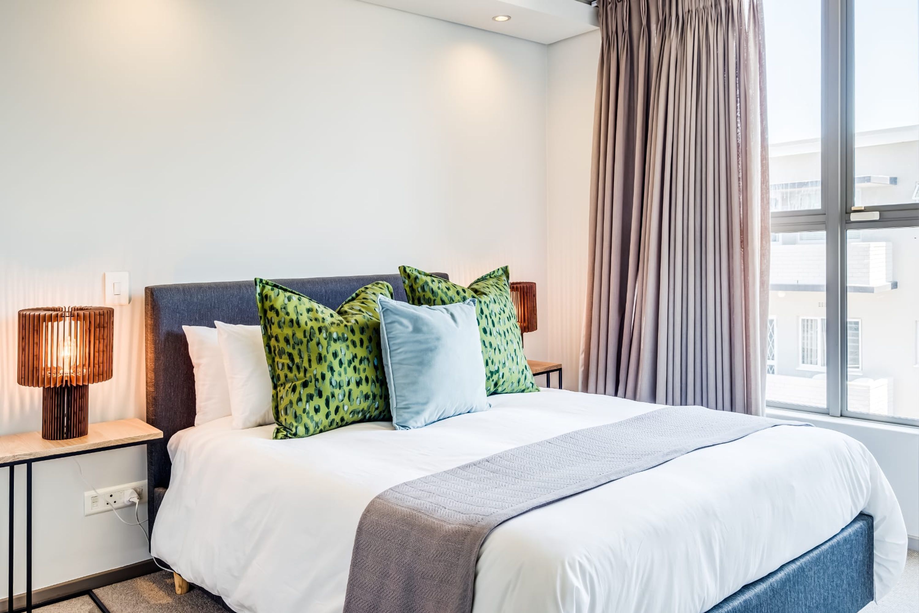 A neatly made bed with white and blue bedding, a grey throw blanket, and green patterned pillows. There is a wooden bedside table with a lamp, a power outlet, and a window with curtains.