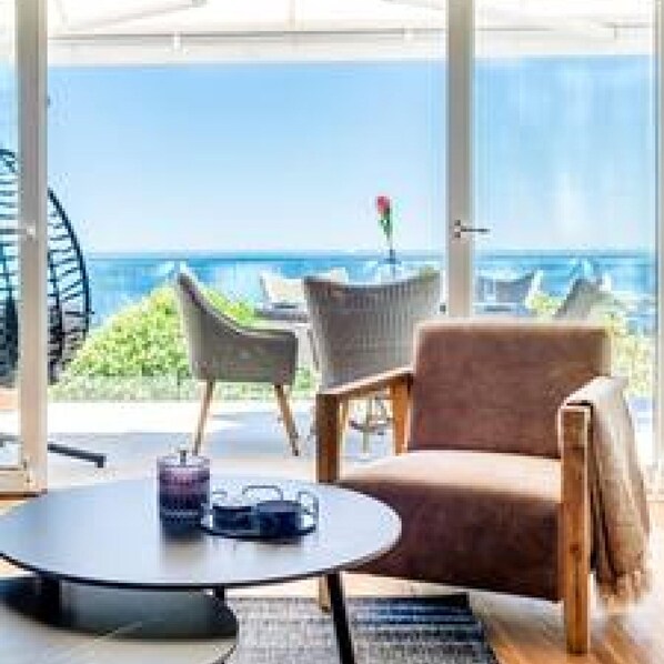 A sunlit room with a large window overlooking the sea, furnished with a round table, two chairs, and a small table with a flower vase.