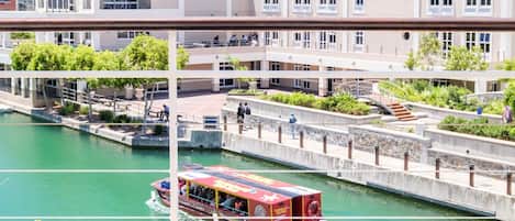 Une vue depuis un balcon donnant sur une rivière, avec un bateau rouge, un immeuble à plusieurs étages et une table sur laquelle se trouvent un magazine et des verres.