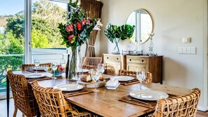 A dining area with a wooden table set for six, surrounded by wicker chairs. A large window offers a view of greenery and hills.