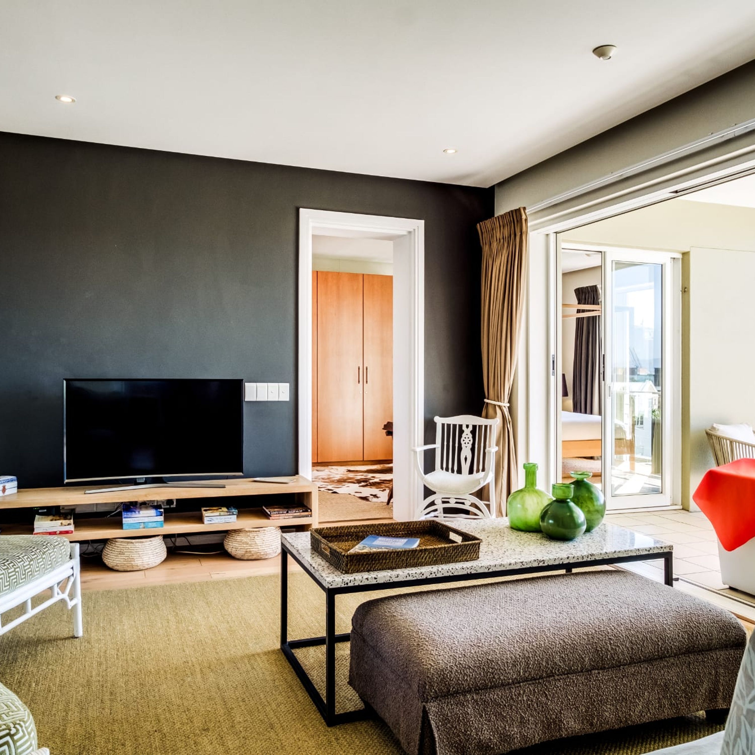 Un salon moderne avec un téléviseur à écran plat, une table basse, un canapé et un fauteuil. Une porte donne accès à un balcon avec vue.