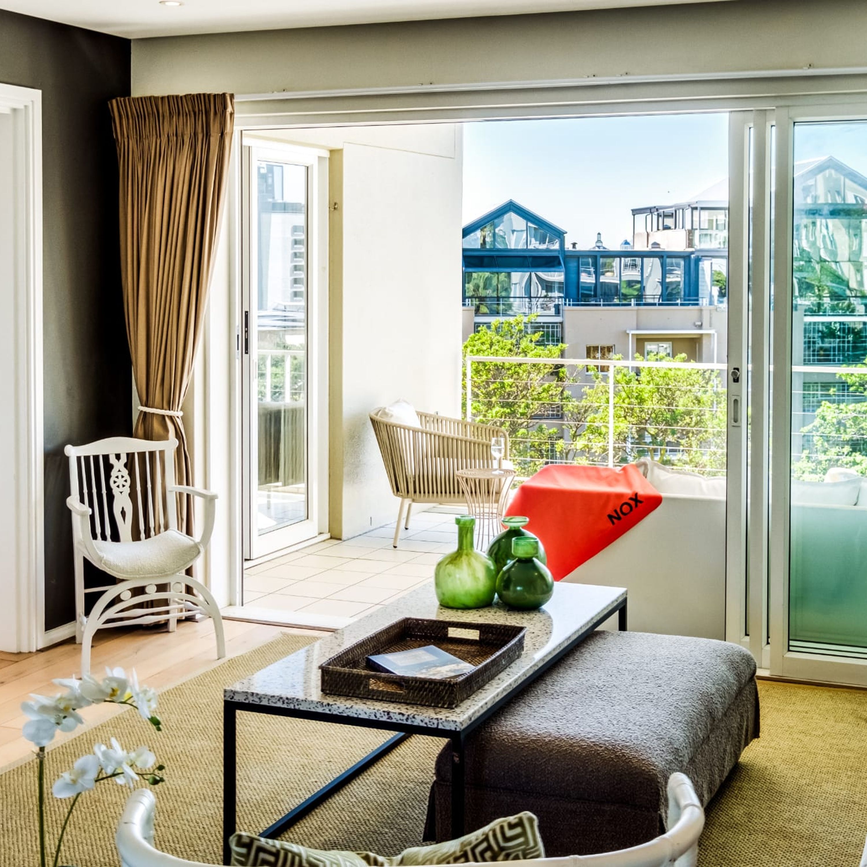 A modern living room with a white chair, a sofa, a glass coffee table, and a view of a building through a glass door.