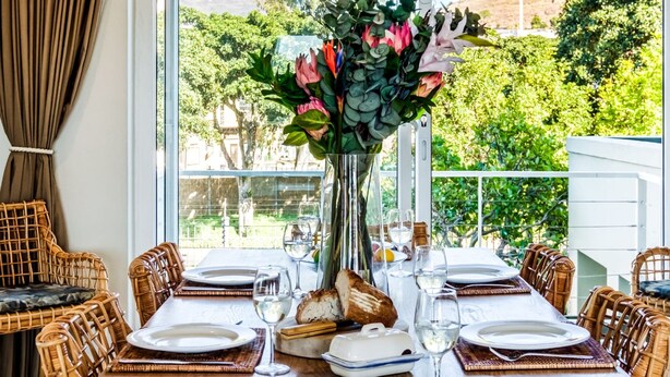 A dining table set for four with wicker chairs, a vase of flowers, and a view of greenery and hills.