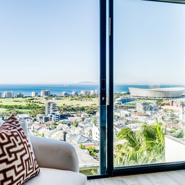 A modern living room with a large window overlooking a coastal cityscape, including a stadium and residential buildings. - Luxurious Villa w Sea City Views Splendor Views (Cape Town)