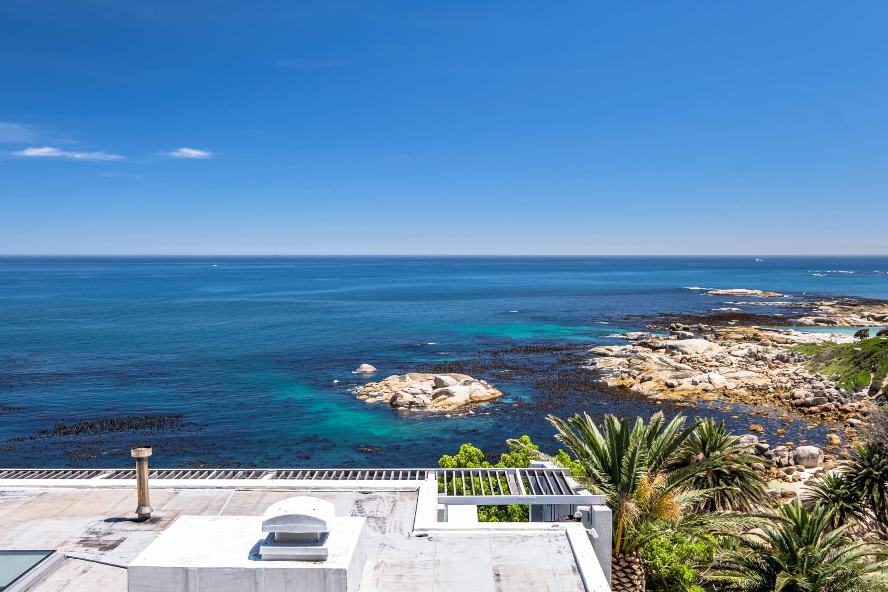 A coastal view with a rocky shoreline, clear blue sea, and a building with a balcony overlooking the ocean.