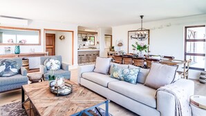 A spacious living room with a sofa, coffee table, and decorative items. A kitchen area is visible in the background with a dining table and chairs. - Luxurious Camps Bay Retreat w Sea Views Rock Residence (Cape Town)