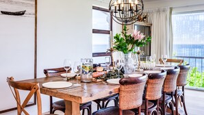 A dining area with a wooden table set for four, surrounded by wicker chairs, a chandelier, and a vase with flowers. - Luxurious Camps Bay Retreat w Sea Views Rock Residence (Cape Town)
