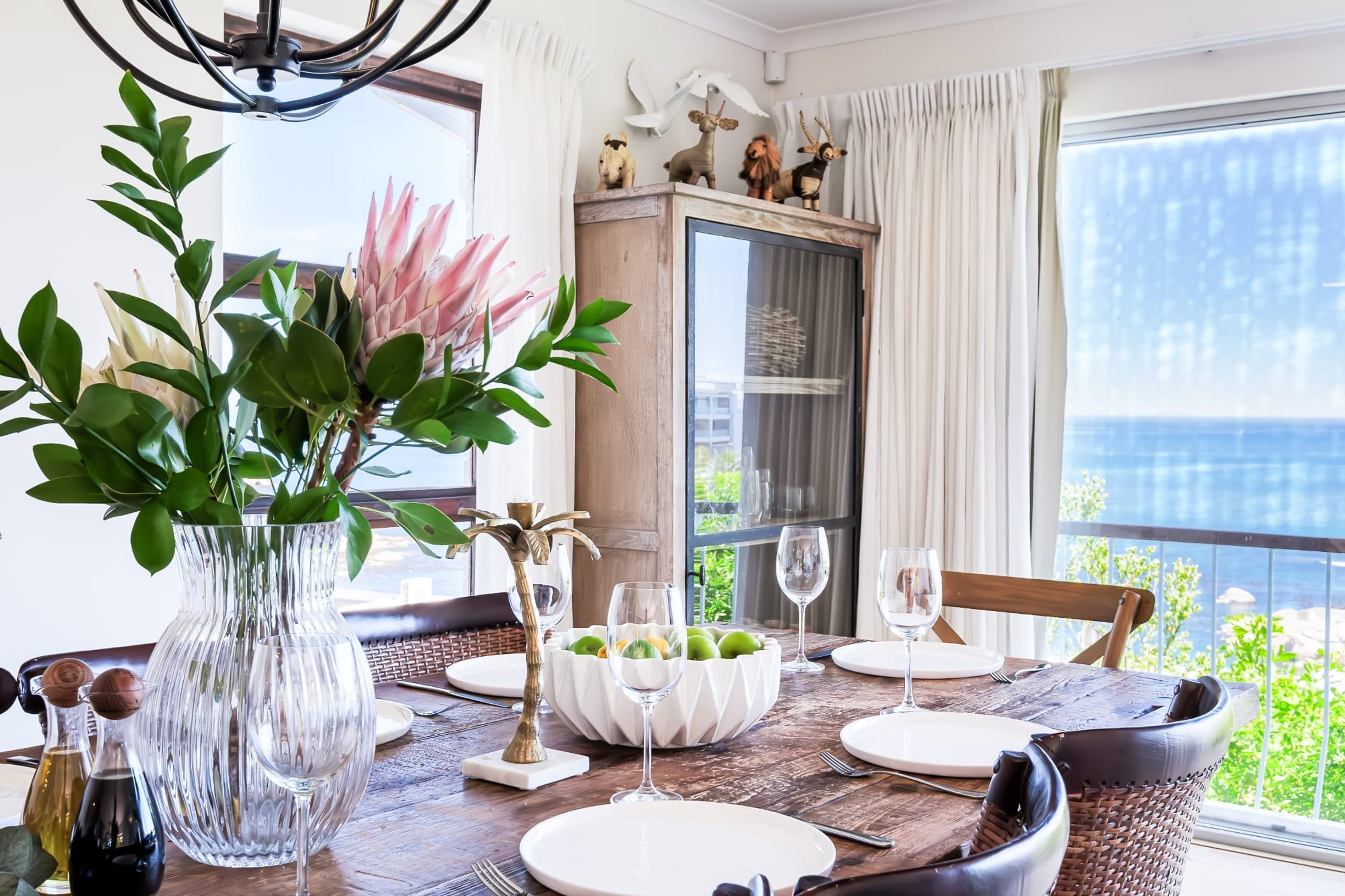 A dining table set with white plates, wine glasses, and a bowl of green apples. There is a vase with green leaves and a pink flower on the table.