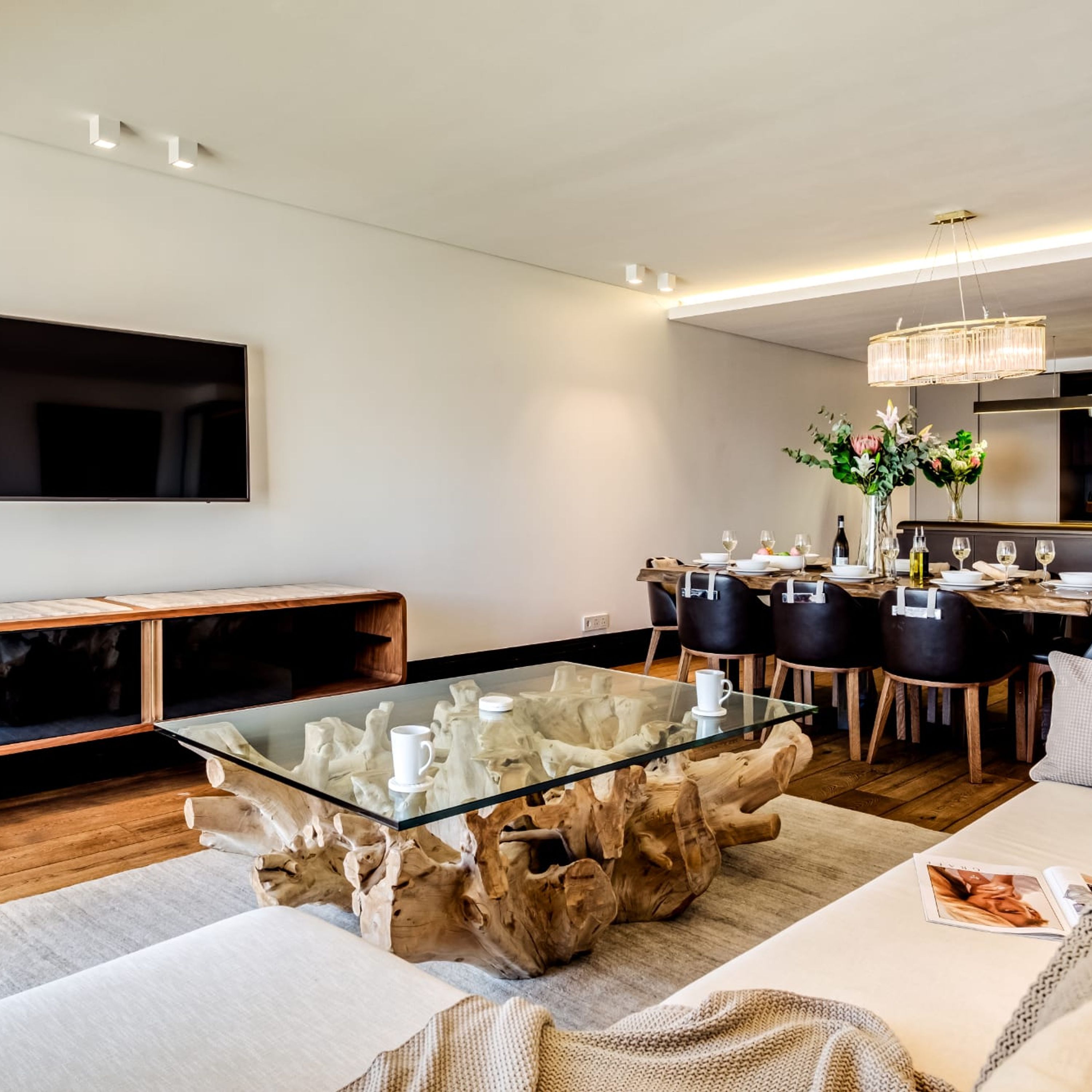 A modern living room with a glass coffee table, wooden accents, a flat-screen TV, and a dining area with a chandelier.