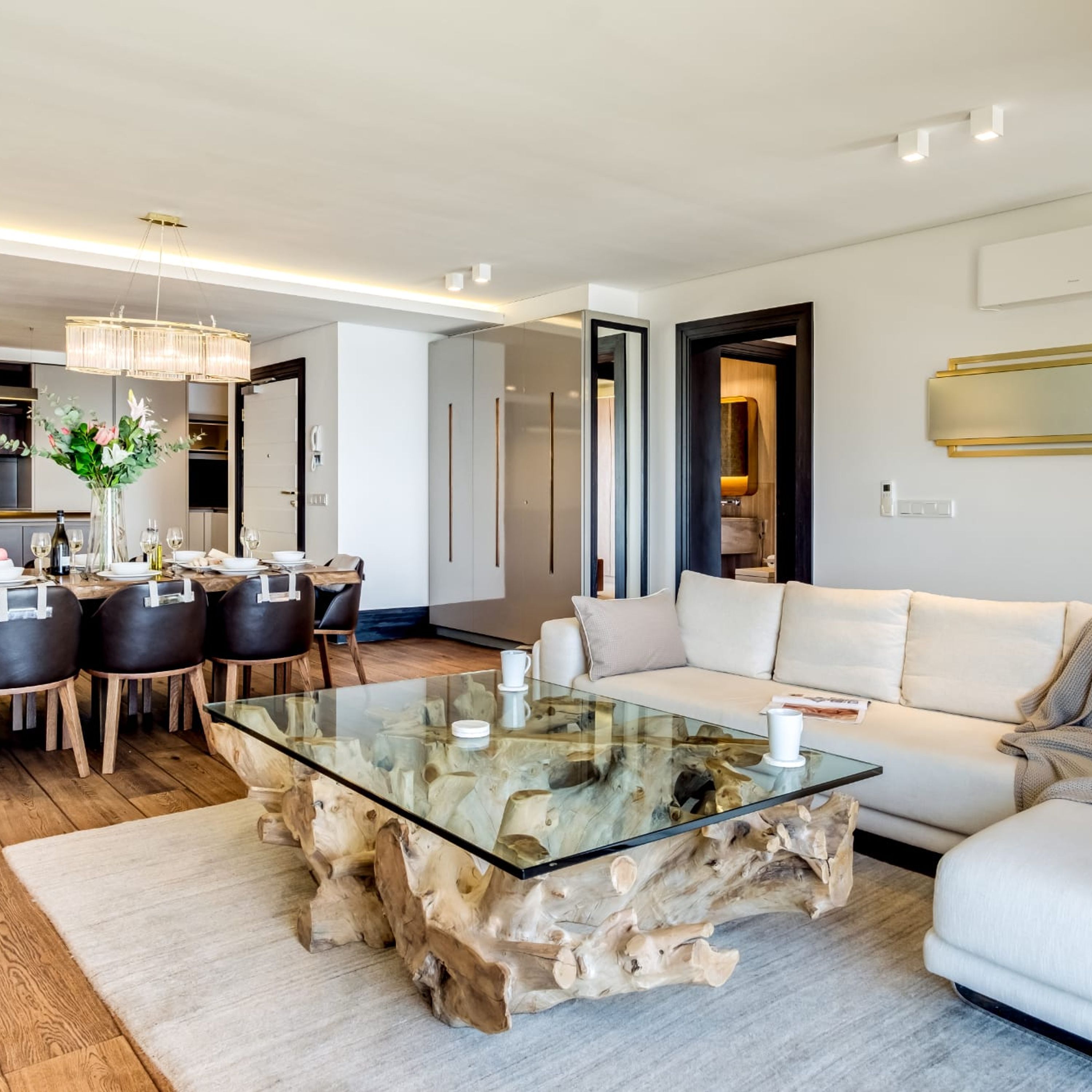 A modern living room with a glass coffee table, a white sofa, and a dining area with a wooden table and chairs.