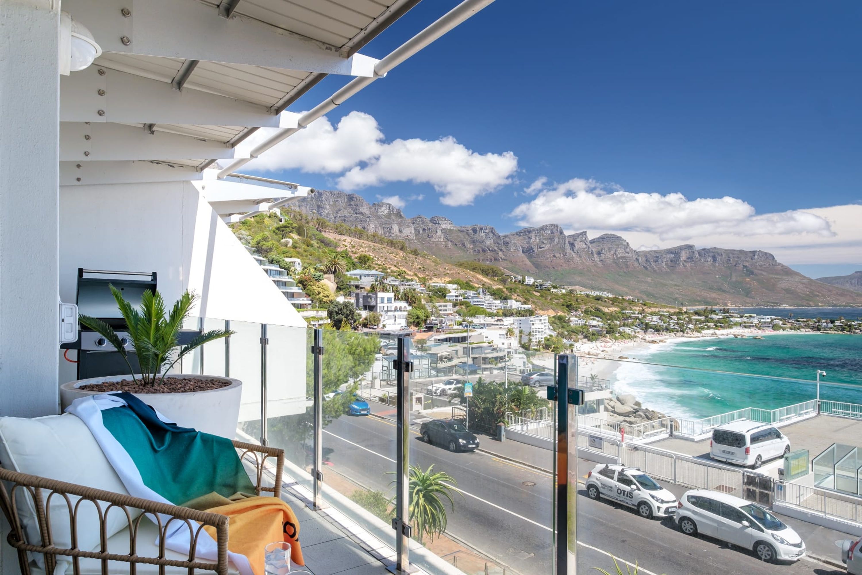 Un balcón con vistas a un pueblo costero, una calle con coches y una cordillera.