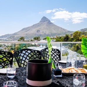 A table with a black mug, a glass of water, a bottle of water, and a plate of food, with a mountain view in the background. - Elegant Apartment w Ocean Views Quenera Apartment (Cape Town)