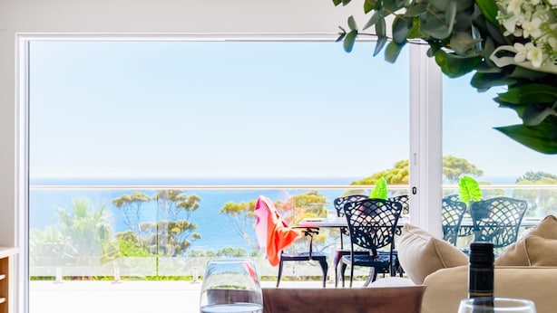 A table with a plate of fruit, a bottle of wine, and a glass of water, overlooking a scenic view of the ocean and greenery. - Elegant Apartment w Ocean Views Quenera Apartment (Cape Town)