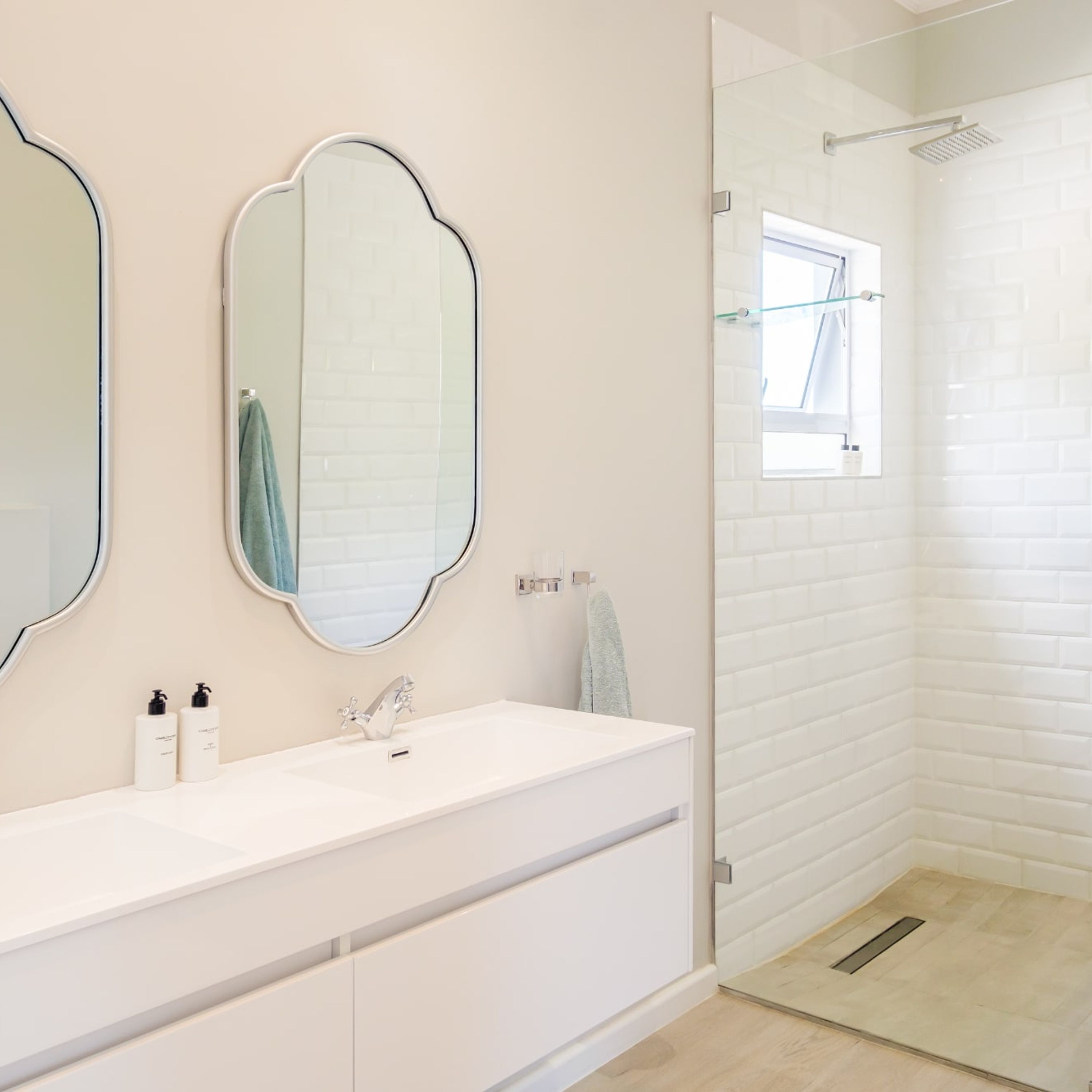 A modern bathroom with a white vanity, two mirrors, a shower with a glass door, and a window.