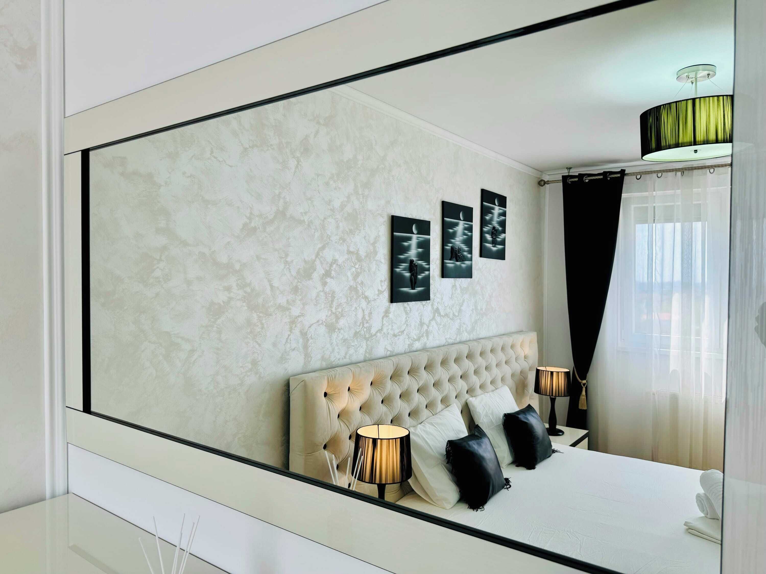 Een moderne slaapkamer met een grote spiegel, een gestoffeerd hoofdeinde, drie ingelijste kunstwerken en twee bedlampen.