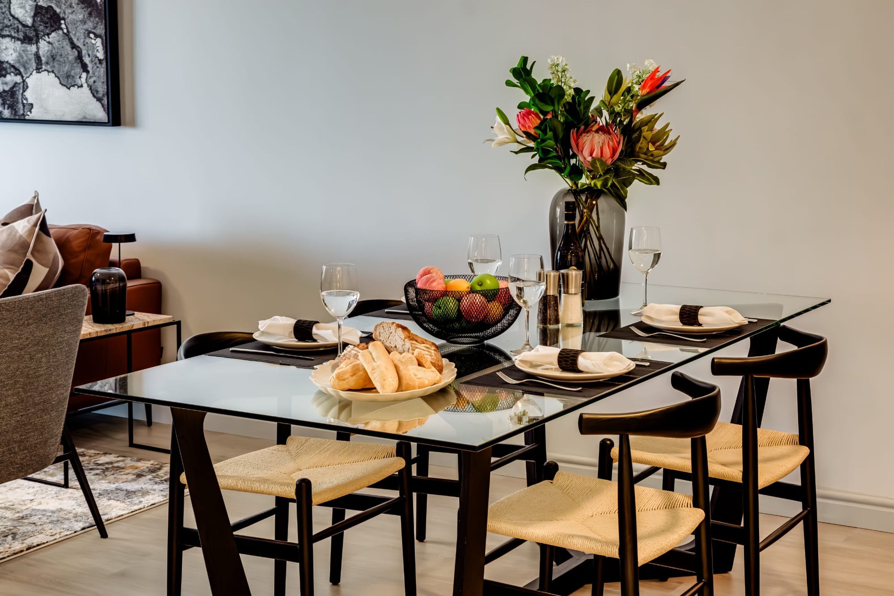 Een eethoek met een glazen tafel, zwarte stoelen, een vaas met bloemen en een schaal met fruit.