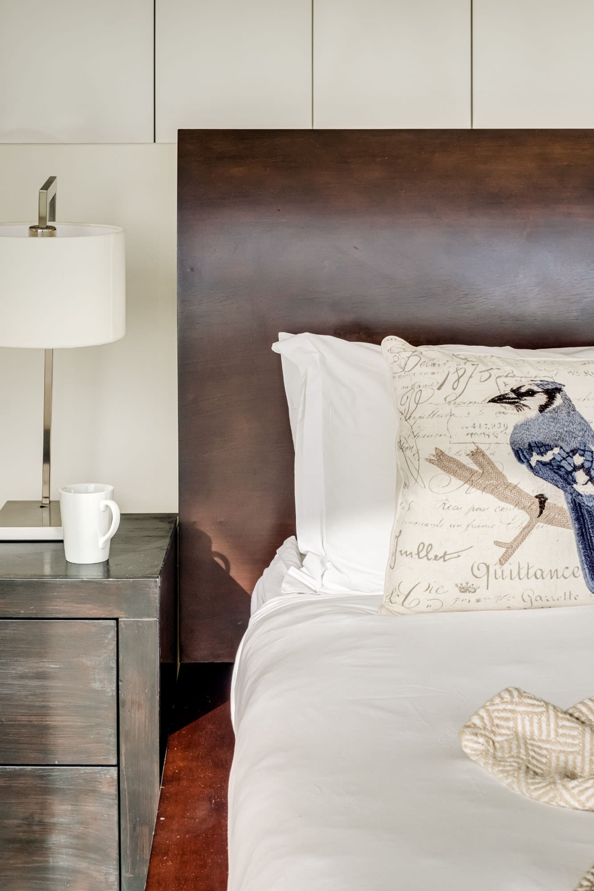 A neatly made bed with a white headboard, a decorative pillow with a bird design, and a bedside table with a lamp and a mug.